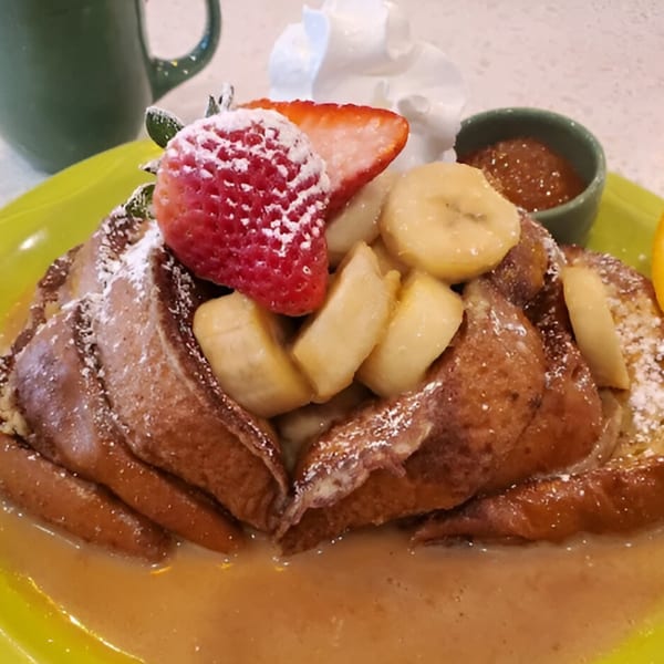 French toast topped with strawberries, bananas, whipped cream, and powdered sugar on a green plate.