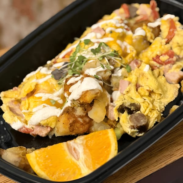 Scrambled eggs with potatoes, ham, and sauce, garnished with microgreens and an orange slice.