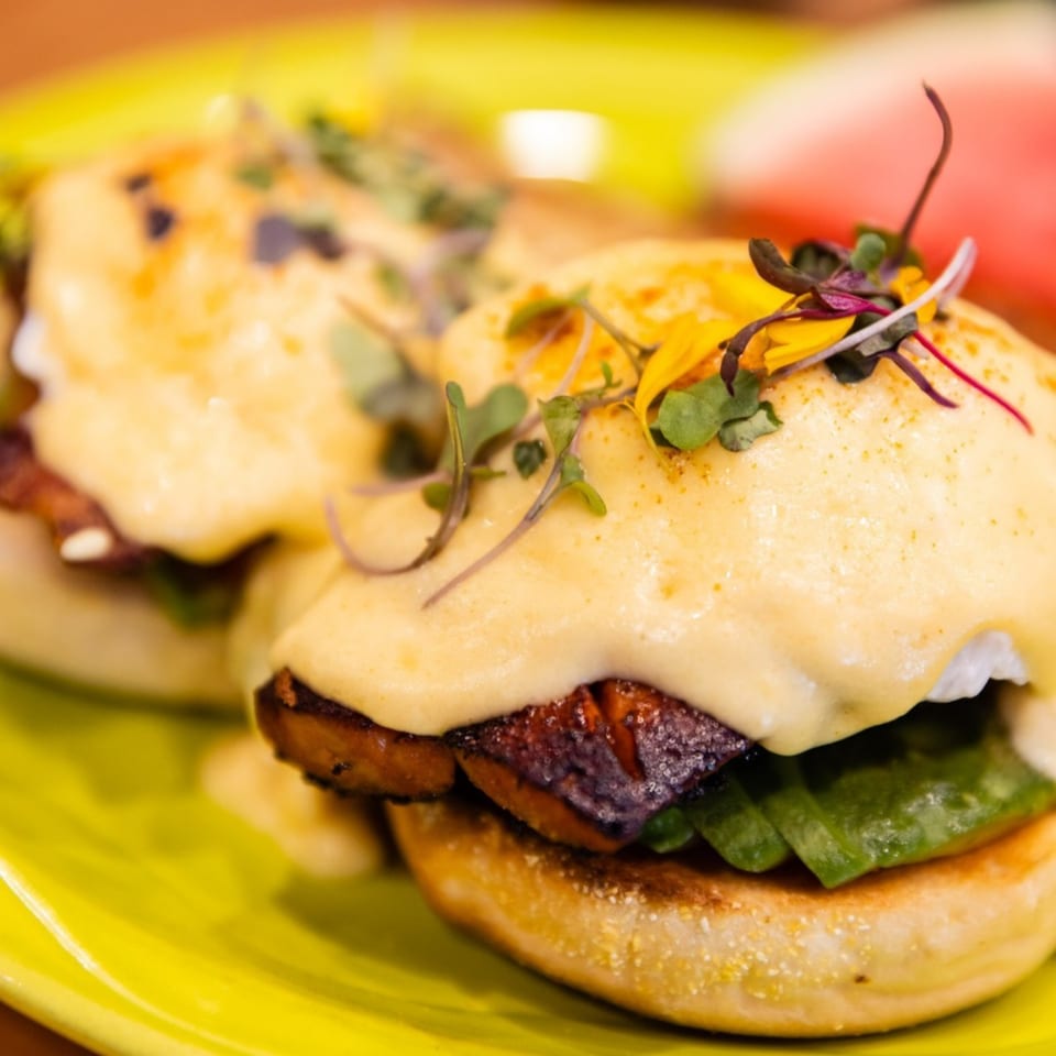 Two servings of eggs Benedict with hollandaise sauce, microgreens, and a side of watermelon.