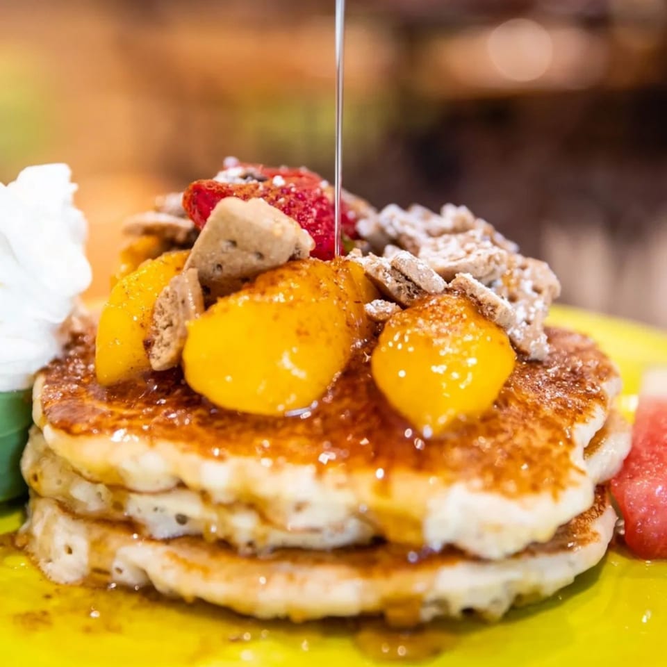Syrup pouring over pancakes topped with peaches, strawberries, cereal, and whipped cream at Lou's Coffee Shop.